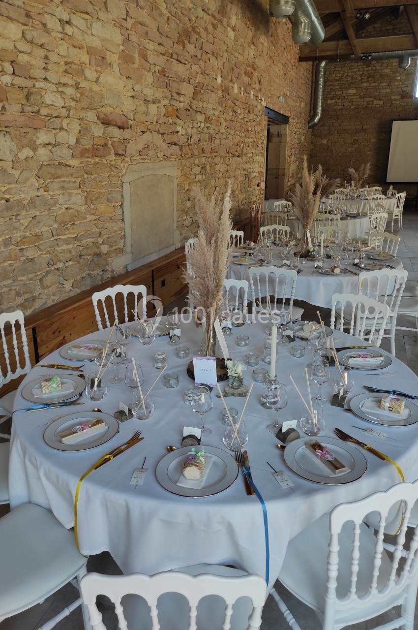 Salle de réception avec tables rondes décorées de nappes blanches, pampas, bougies et vaisselle élégante.