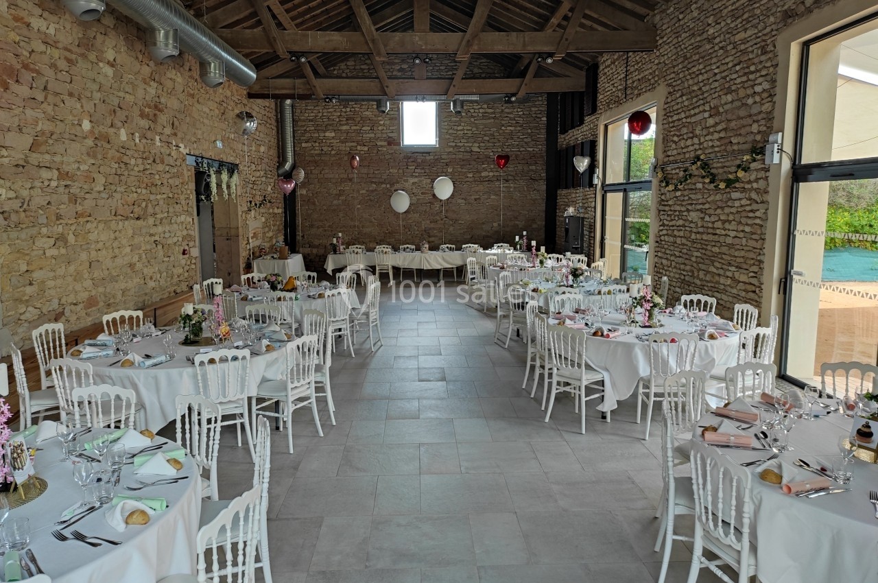Salle de réception en pierre avec tables rondes dressées, décorations florales et lumière naturelle entrant par les fenêtres.