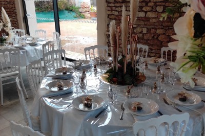 Cour d'une maison en pierre avec terrasse fleurie, arbres, guirlandes lumineuses et sol en gravier.