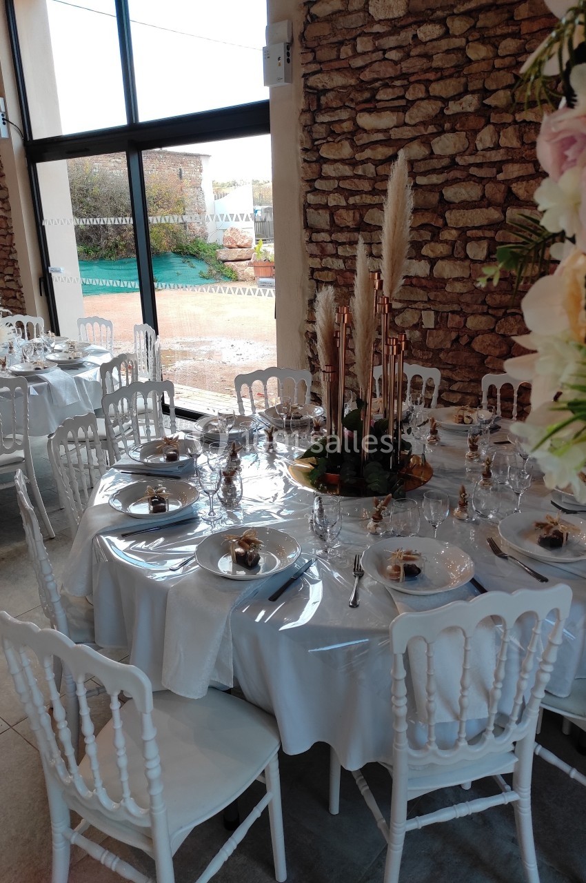 Salle de réception avec tables rondes dressées, nappes blanches, chaises blanches et décoration florale élégante.