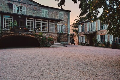 Cour d'une maison en pierre avec terrasse fleurie, arbres, guirlandes lumineuses et sol en gravier.