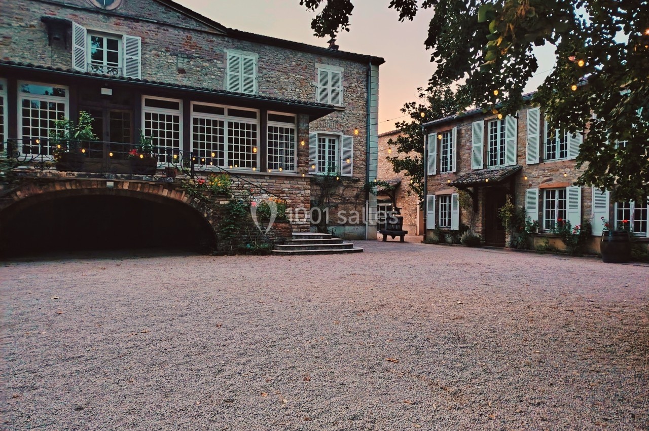 Cour pavée entourée de deux bâtiments en pierre avec volets blancs, éclairée par une lumière douce au crépuscule.