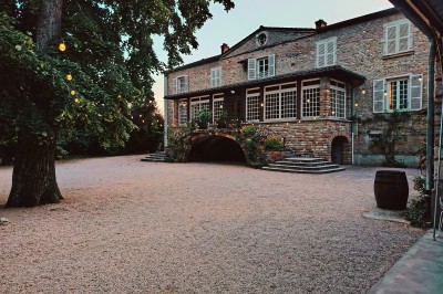 Cour d'une maison en pierre avec terrasse fleurie, arbres, guirlandes lumineuses et sol en gravier.