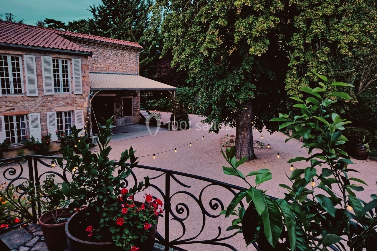 Cour d'une maison en pierre avec terrasse fleurie, arbres, guirlandes lumineuses et sol en gravier.