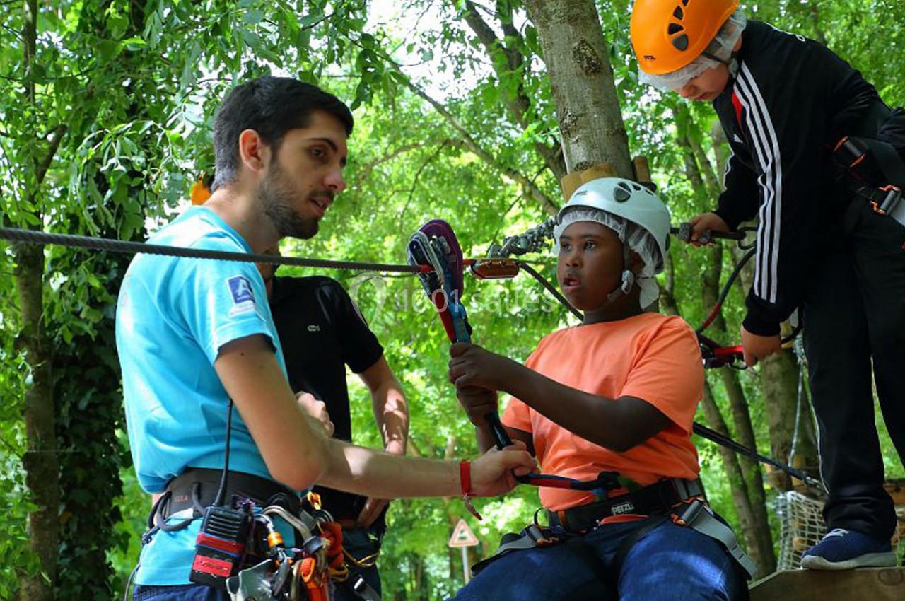 Un instructeur aide un jeune à attacher son harnais lors d'une activité d'accrobranche en forêt.