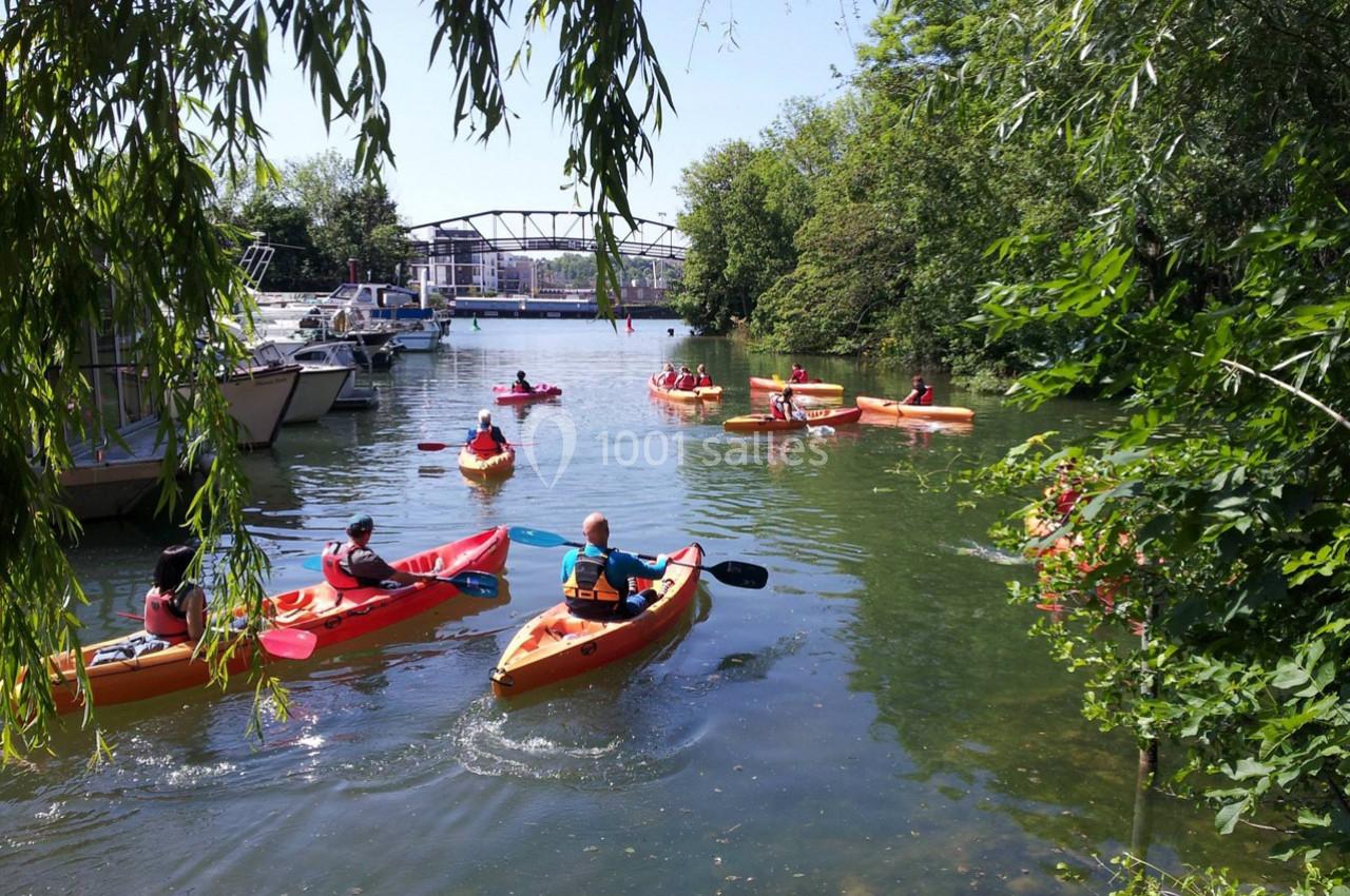 Des personnes pagayant en kayak sur une rivière bordée de végétation, avec des bateaux amarrés et un pont en arrière-plan.