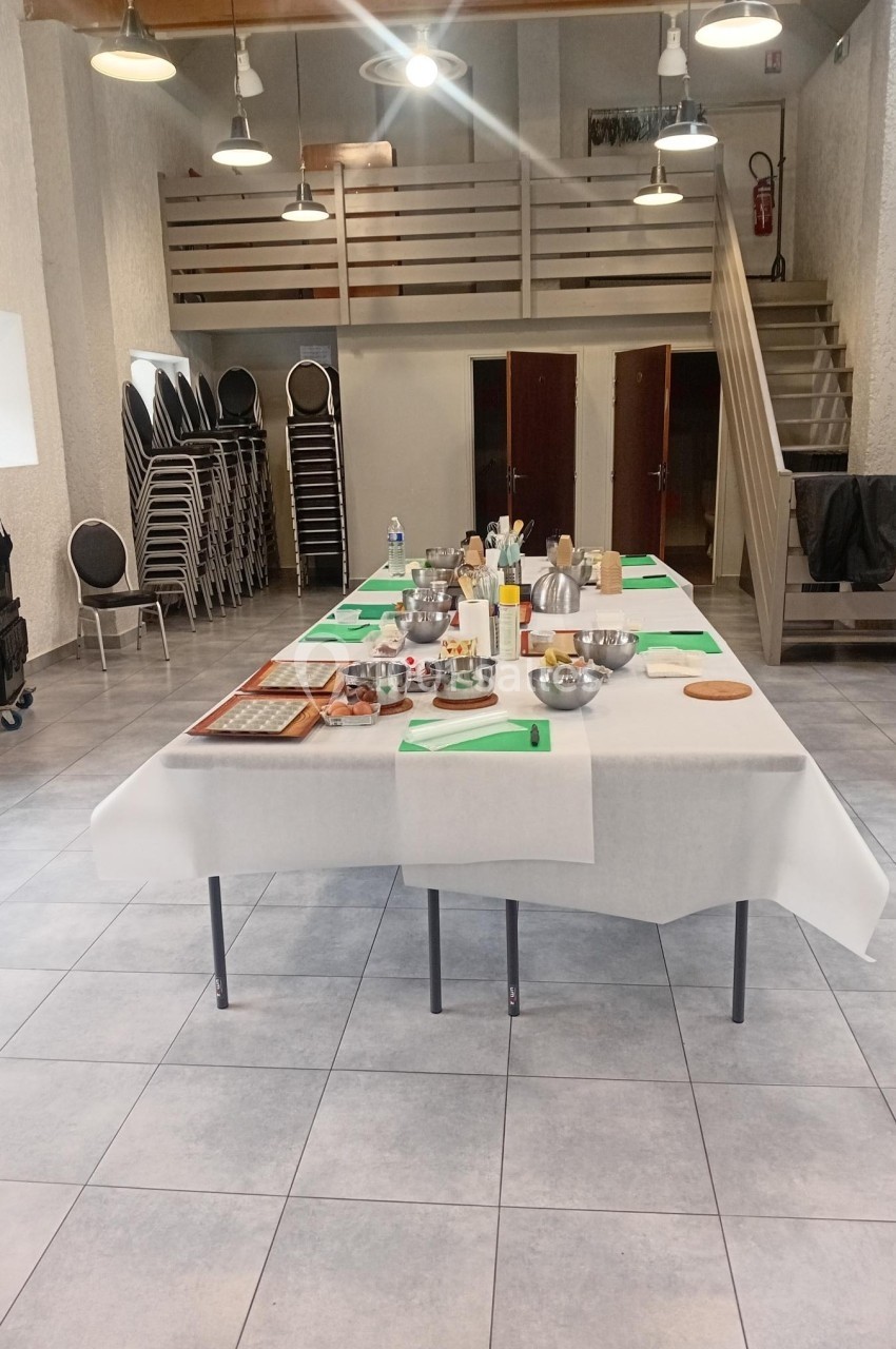 Tables dressées avec des ustensiles de cuisine et ingrédients dans une grande salle équipée de chaises empilées.