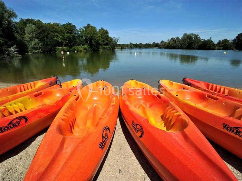 Six kayaks orange alignés sur une plage de sable au bord d'un lac entouré de végétation sous un ciel dégagé.