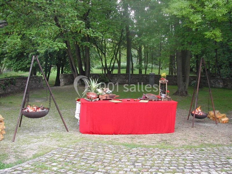 Table drapée de rouge avec des plats disposés, entourée de braseros allumés dans un jardin arboré.