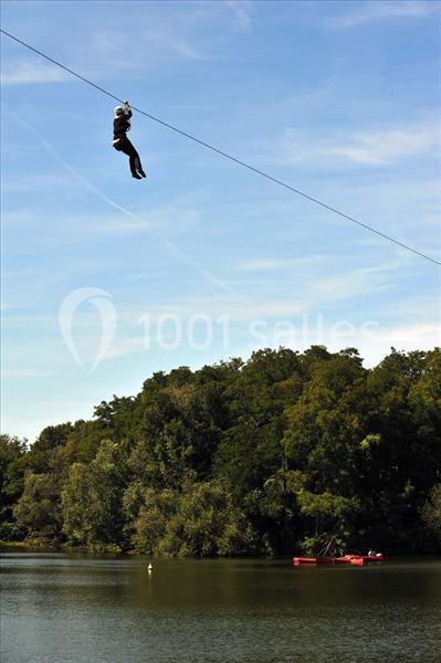Une personne traverse un lac en tyrolienne, avec des arbres en arrière-plan et une barque rouge sur l'eau.