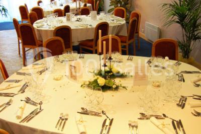 Salle de réception avec tables rondes dressées, nappes blanches, chaises rouges et décorations de ballons colorés.