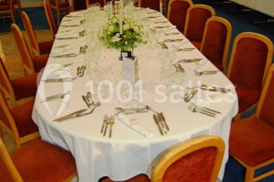 Salle de réception avec tables rondes dressées, nappes blanches, chaises rouges et décorations de ballons colorés.