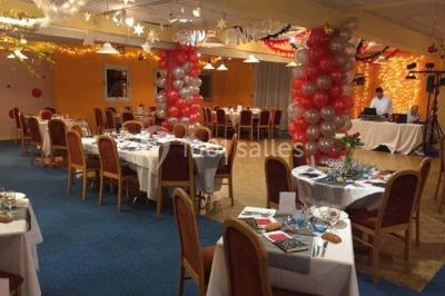 Salle de réception avec tables rondes dressées, nappes blanches, chaises rouges et décorations de ballons colorés.