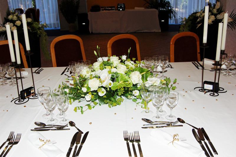 Table dressée avec nappes blanches, centre de table floral blanc et vert, couverts, verres et chandeliers.