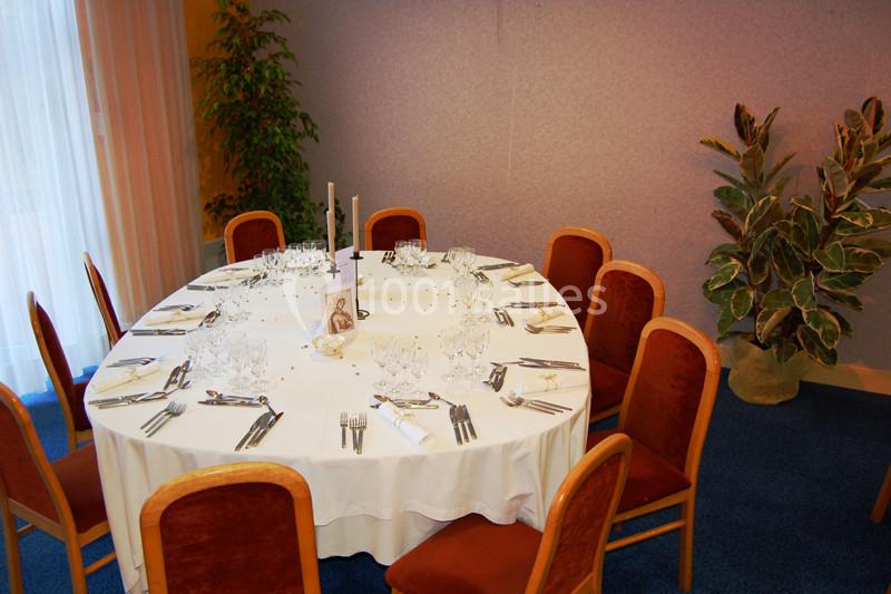 Table ronde dressée avec nappes blanches, verres, couverts et bougies, entourée de chaises en bois avec assises rouges.