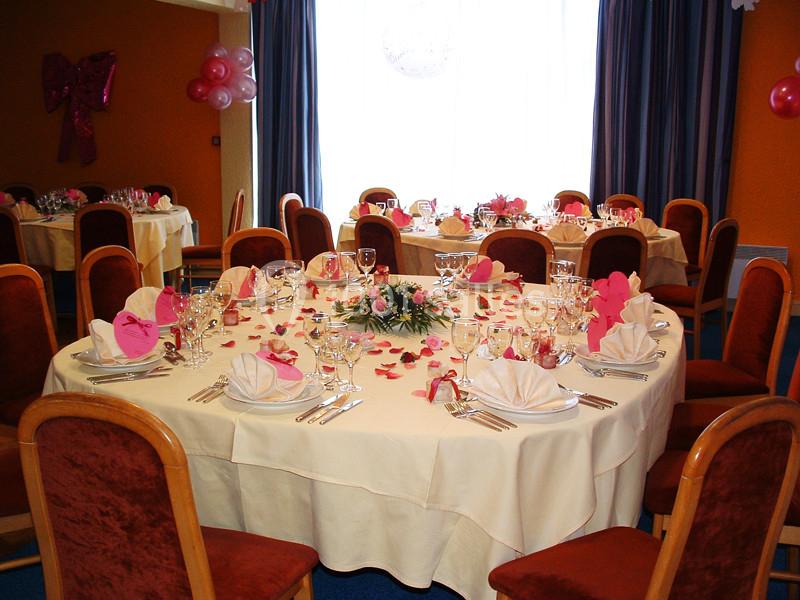 Salle décorée pour un événement avec des tables rondes dressées, nappes blanches, serviettes pliées et pétales roses.