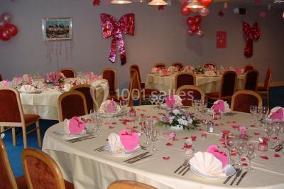 Salle de réception avec tables rondes dressées, nappes blanches, chaises rouges et décorations de ballons colorés.