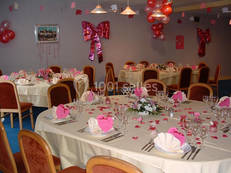 Salle décorée pour une réception avec tables rondes dressées, nappes blanches, décorations roses et ballons suspendus.