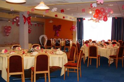 Salle de réception avec tables rondes dressées, nappes blanches, chaises rouges et décorations de ballons colorés.