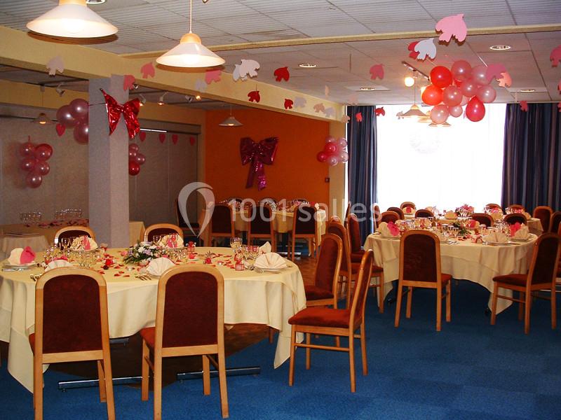 Salle décorée pour une réception avec tables rondes dressées, chaises en bois et ballons roses suspendus.