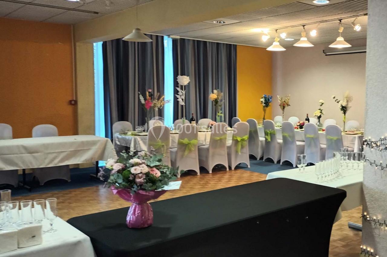 Salle décorée pour un événement avec tables dressées, chaises ornées de nœuds verts et fleurs en centre de table.