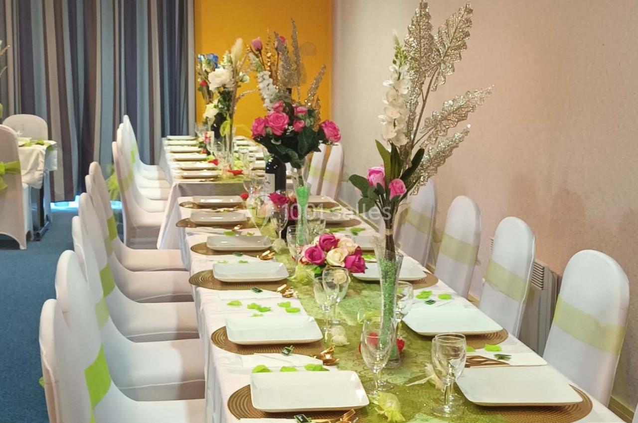Table dressée avec nappes vertes, chaises blanches, vaisselle blanche et décorations florales colorées.
