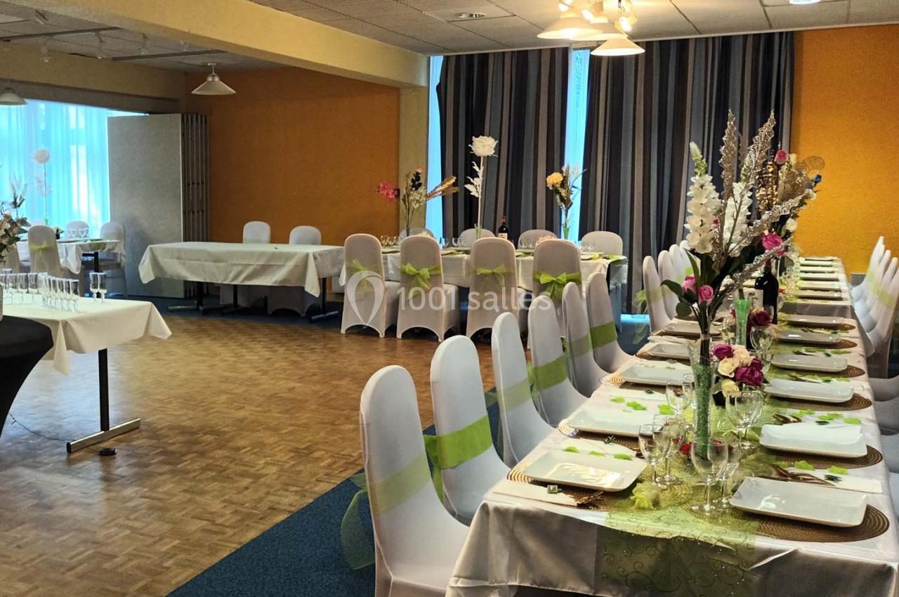 Salle de réception décorée avec des tables dressées, nappes blanches, chaises ornées de rubans verts et arrangements floraux.