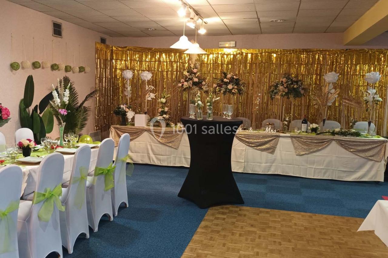 Salle décorée pour une réception avec tables dressées, chaises blanches ornées de nœuds verts et fond doré fleuri.
