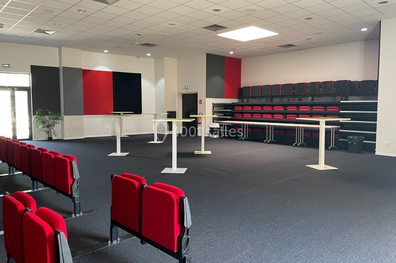 Salle de conférence vide avec sièges rouges, tables hautes blanches et gradins en arrière-plan.