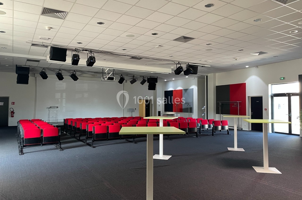 Salle de conférence moderne avec rangées de sièges rouges, pupitres blancs et éclairage suspendu.