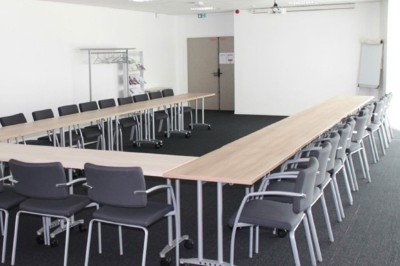 Salle de réunion lumineuse avec des tables disposées en U, des chaises grises et un tableau blanc au fond.