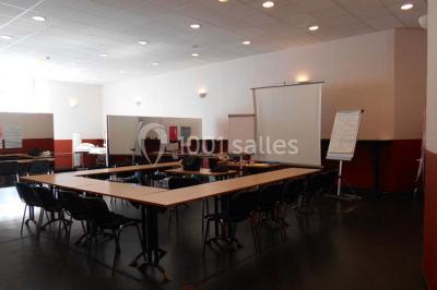 Salle vide avec sol noir, murs blancs et gris, éclairage au plafond, plante en pot et porte métallique au fond.