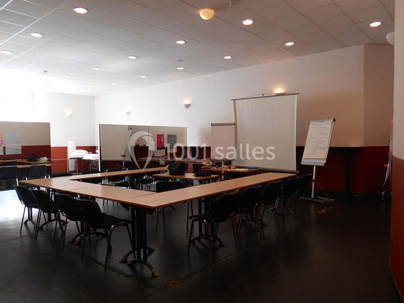 Salle de réunion avec tables disposées en U, chaises, tableau blanc, écran de projection et paperboards.