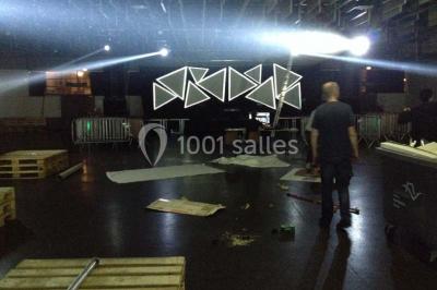 Salle vide avec sol noir, murs blancs et gris, éclairage au plafond, plante en pot et porte métallique au fond.