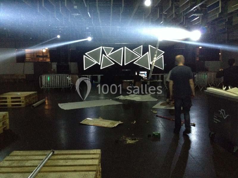 Salle en préparation avec des structures géométriques lumineuses, palettes au sol et quelques personnes présentes.