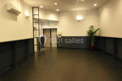 Salle vide avec sol noir, murs blancs et gris, éclairage au plafond, plante en pot et porte métallique au fond.