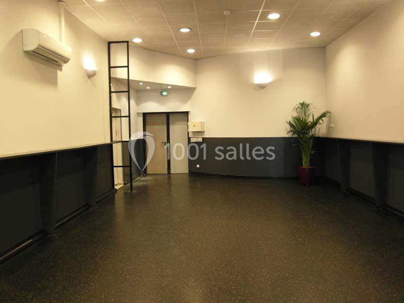 Salle vide avec sol noir, murs blancs et gris, éclairage au plafond, plante en pot et porte métallique au fond.