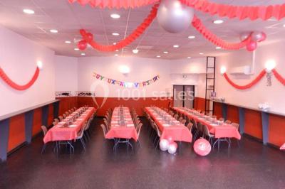 Salle vide avec sol noir, murs blancs et gris, éclairage au plafond, plante en pot et porte métallique au fond.