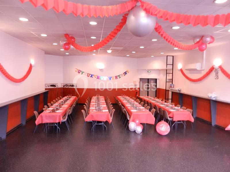 Salle décorée pour un anniversaire avec tables alignées, nappes rouges, guirlandes et ballons suspendus.