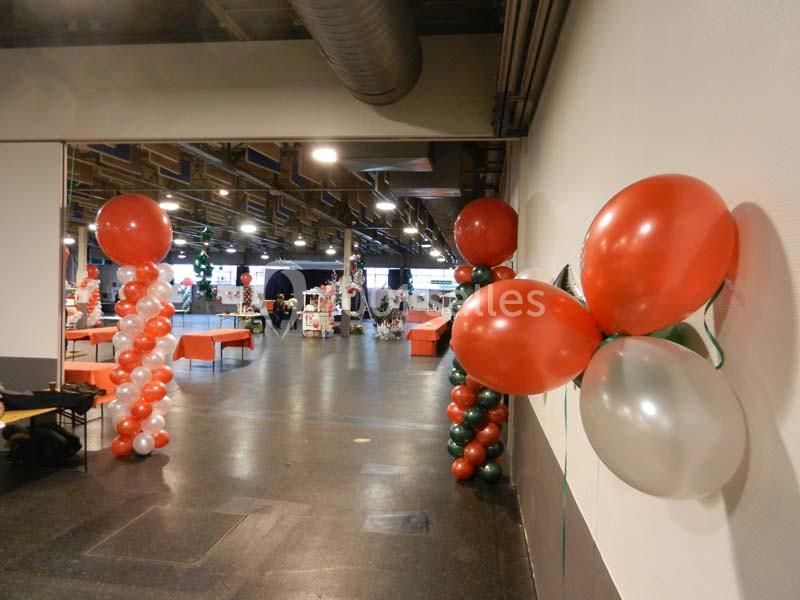 Salle décorée avec des ballons rouges, blancs et verts, tables et stands visibles en arrière-plan.