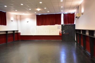 Salle vide avec sol noir, murs blancs et gris, éclairage au plafond, plante en pot et porte métallique au fond.