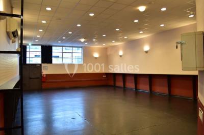 Salle vide avec sol noir, murs blancs et gris, éclairage au plafond, plante en pot et porte métallique au fond.