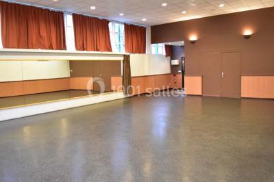 Salle vide avec sol noir, murs blancs et gris, éclairage au plafond, plante en pot et porte métallique au fond.