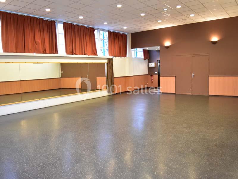 Salle vide avec sol en lino, murs partiellement recouverts de bois, miroir mural et rideaux rouges aux fenêtres.