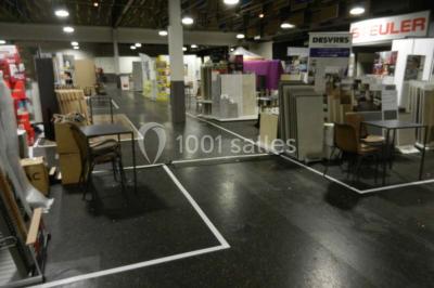 Salle vide avec sol noir, murs blancs et gris, éclairage au plafond, plante en pot et porte métallique au fond.