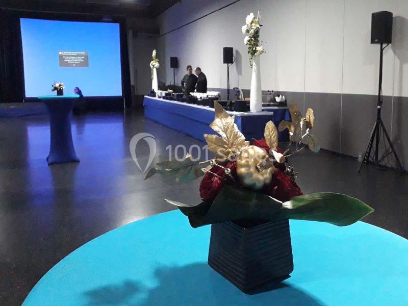 Arrangement floral sur une table bleue au premier plan, avec une salle de réception et un écran en arrière-plan.