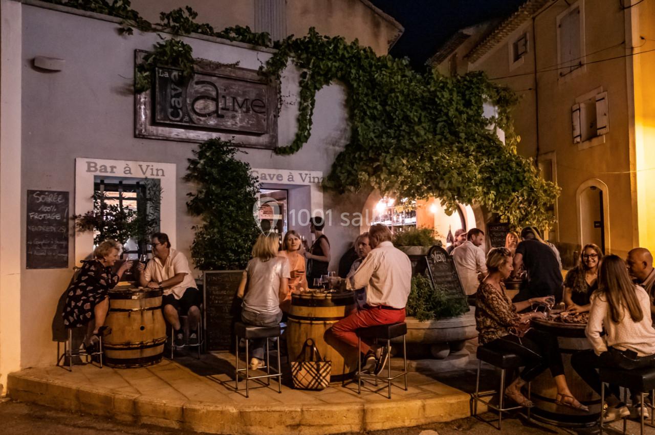 Terrasse animée d'un bar à vin en soirée, avec des clients attablés autour de tonneaux et une façade couverte de végétation.