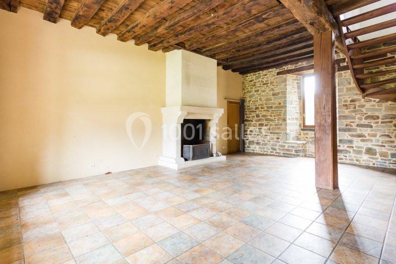 Salle vide avec cheminée en pierre, murs en pierre et plâtre, plafond en poutres apparentes et sol en carrelage.