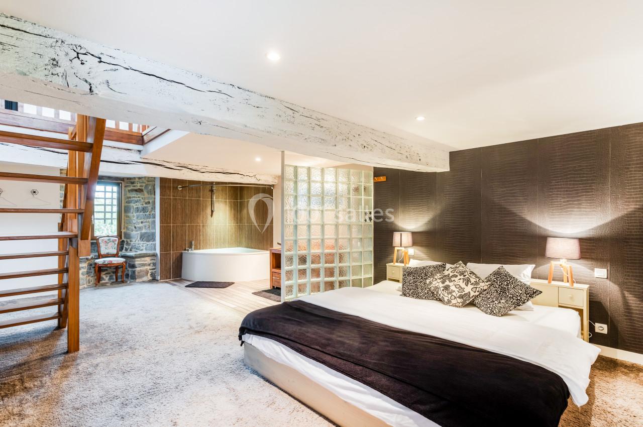 Chambre spacieuse avec lit double, poutres apparentes, baignoire en fond et escalier en bois menant à un étage supérieur.