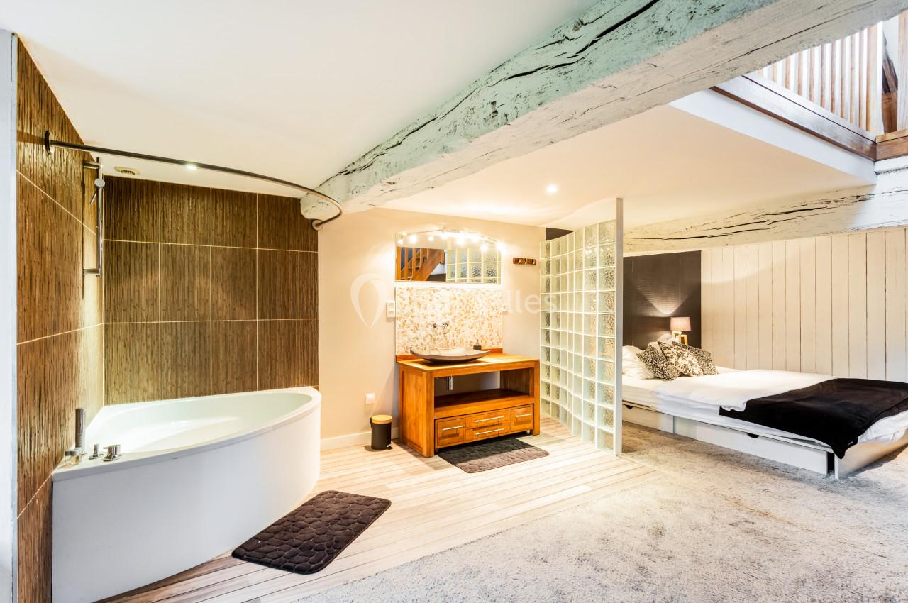 Salle de bain ouverte avec baignoire d'angle, vasque en bois, paroi en verre et lit dans un espace attenant.